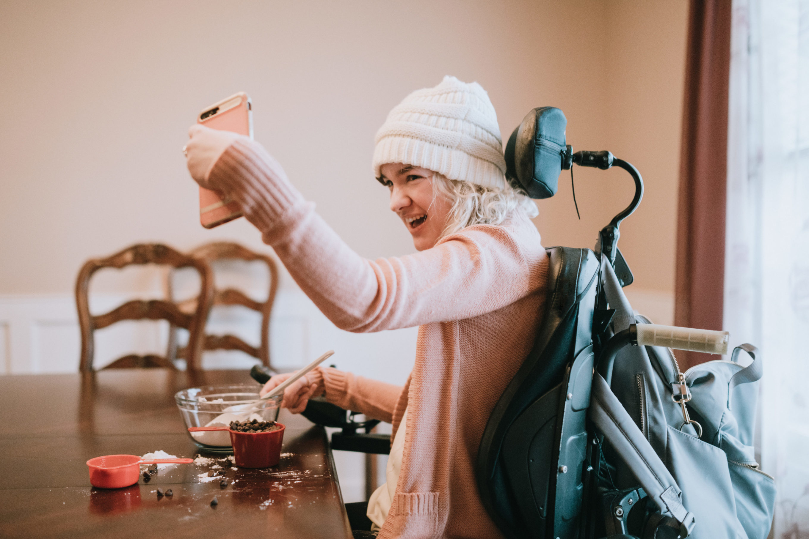 An independent young adult woman with cerebral palsy going about some of her daily routines at home.  She smiles for a self portrait with her smartphone while mixing together ingredients for making chocolate cookies.