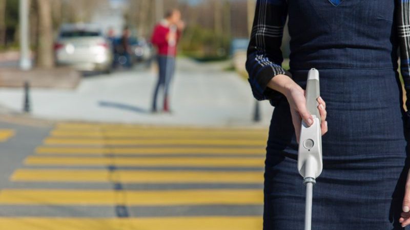 A woman in a blue dress crosses a street using a smart cane device.